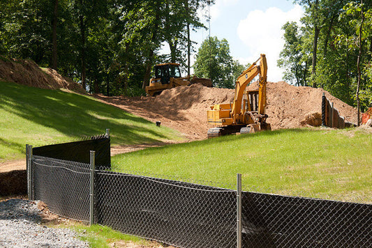 Stormwater Sediment Control BMP Silt Fence with chainlink fence reinforcement. Excavator and bulldozer on a construction site establishing 70 percent pre construction vegetation. Germinating with green grass and per-existing trees in the background. Final stabilization to achieve CGP compliance to file Notice of Intent (NOT)